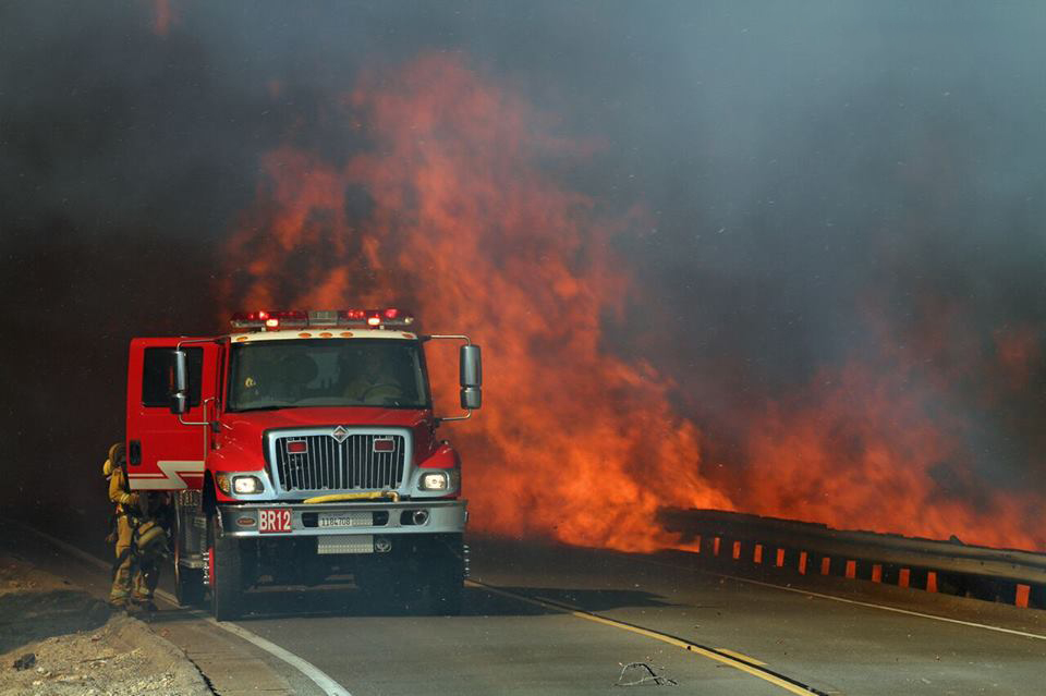 Fire engine and crew working directly beside flames along a road